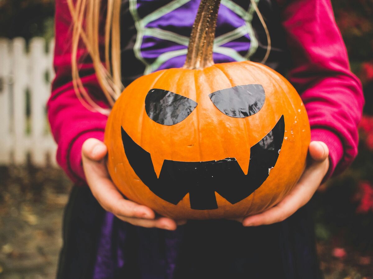 A person in costume holds a pumpkin with a painted black jack-o'-lantern face, set against an outdoor background with a white fence.