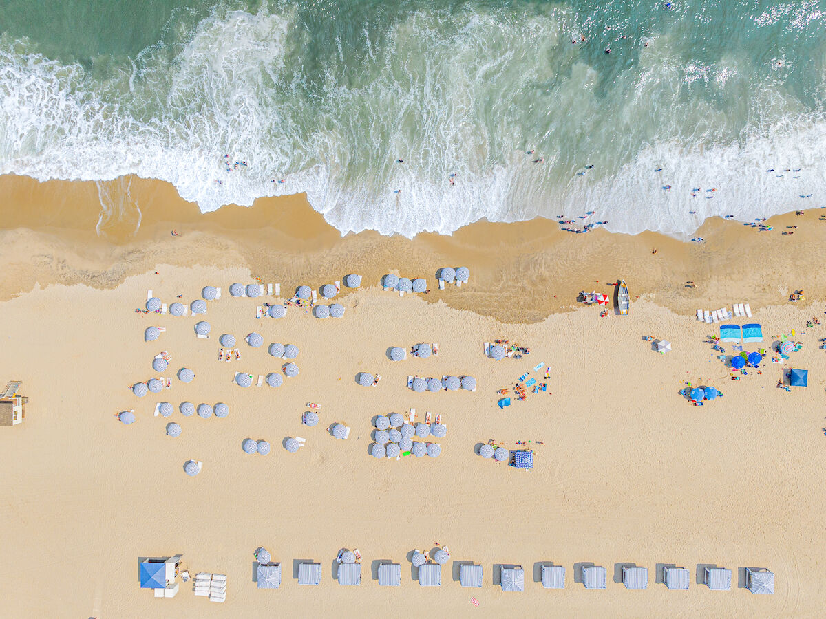 Aerial view of a beach showing umbrellas, people, and waves crashing on the shore, creating a vibrant, summery scene.