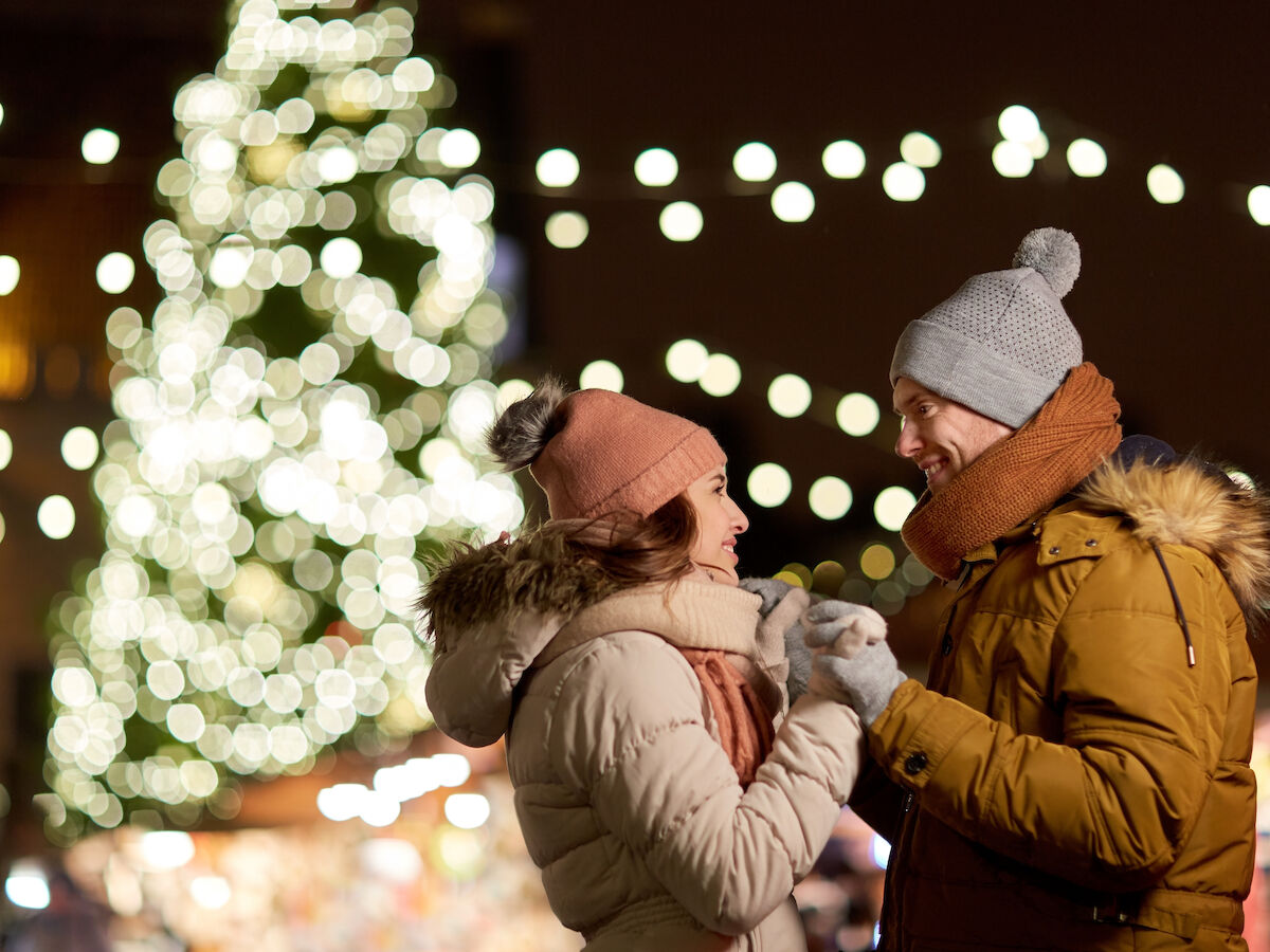 A couple in winter clothing stands close together, smiling under festive lights with a blurred, lit Christmas tree in the background.