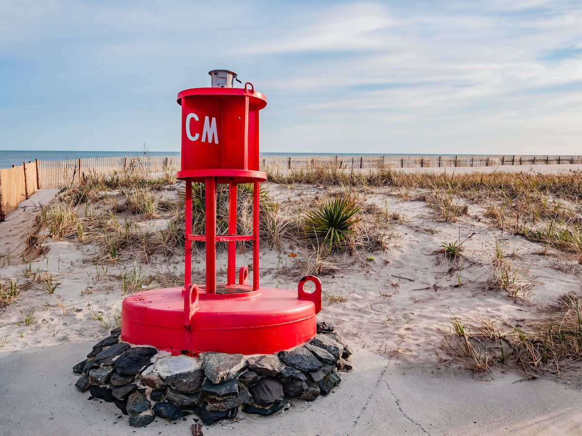 A red buoy marked "CM" is placed on a sandy beach surrounded by rocks and dune grass under a partly cloudy sky.
