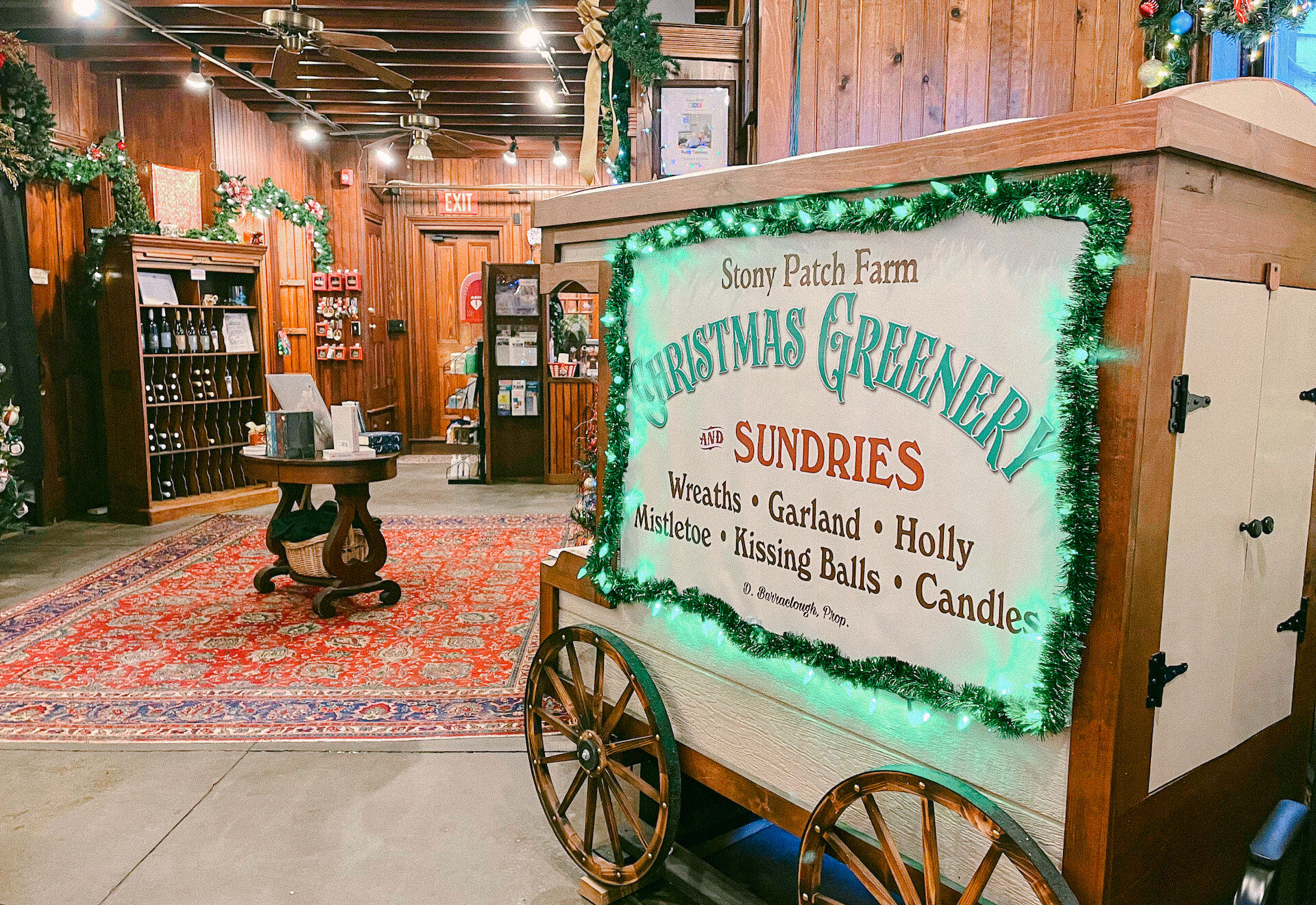 A festive shop interior with a cart displaying "Christmas Greenery" items like wreaths and mistletoe. Warm lighting and decorations abound.