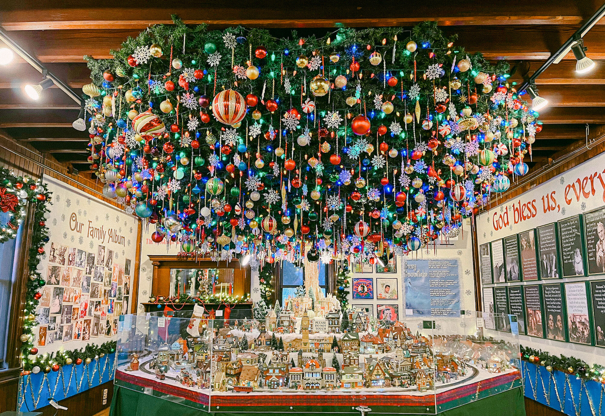 The image shows an indoor festive display with a decorated upside-down Christmas tree and a miniature village scene underneath it.