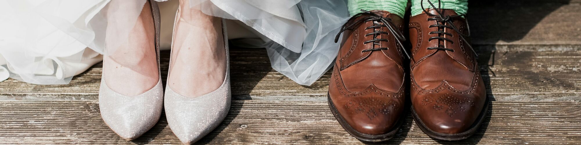 A pair of wedding shoes and brown dress shoes with green socks on a wooden deck.