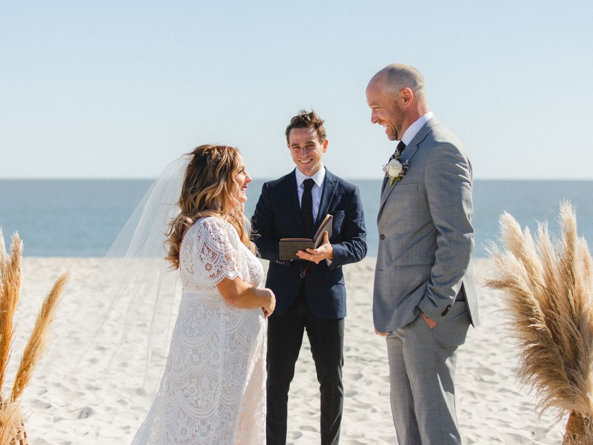 A couple is getting married on a beach, with a blue ocean in the background and pampas grass decorations on either side, officiated by a man in a suit.