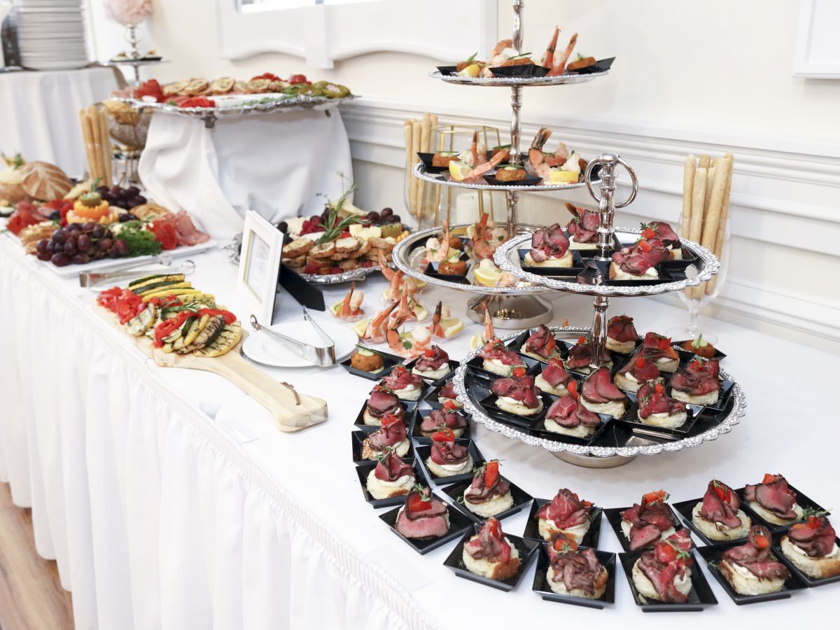 A table set up with an assortment of appetizers, including fruit, meat, and sandwich bites, arranged elegantly for what appears to be a buffet.