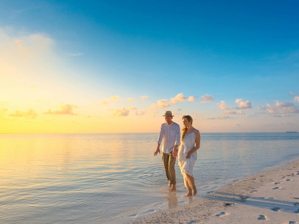 A couple walks along a beach at sunset, with the sun setting over calm waters and a clear sky, creating a serene and romantic atmosphere.