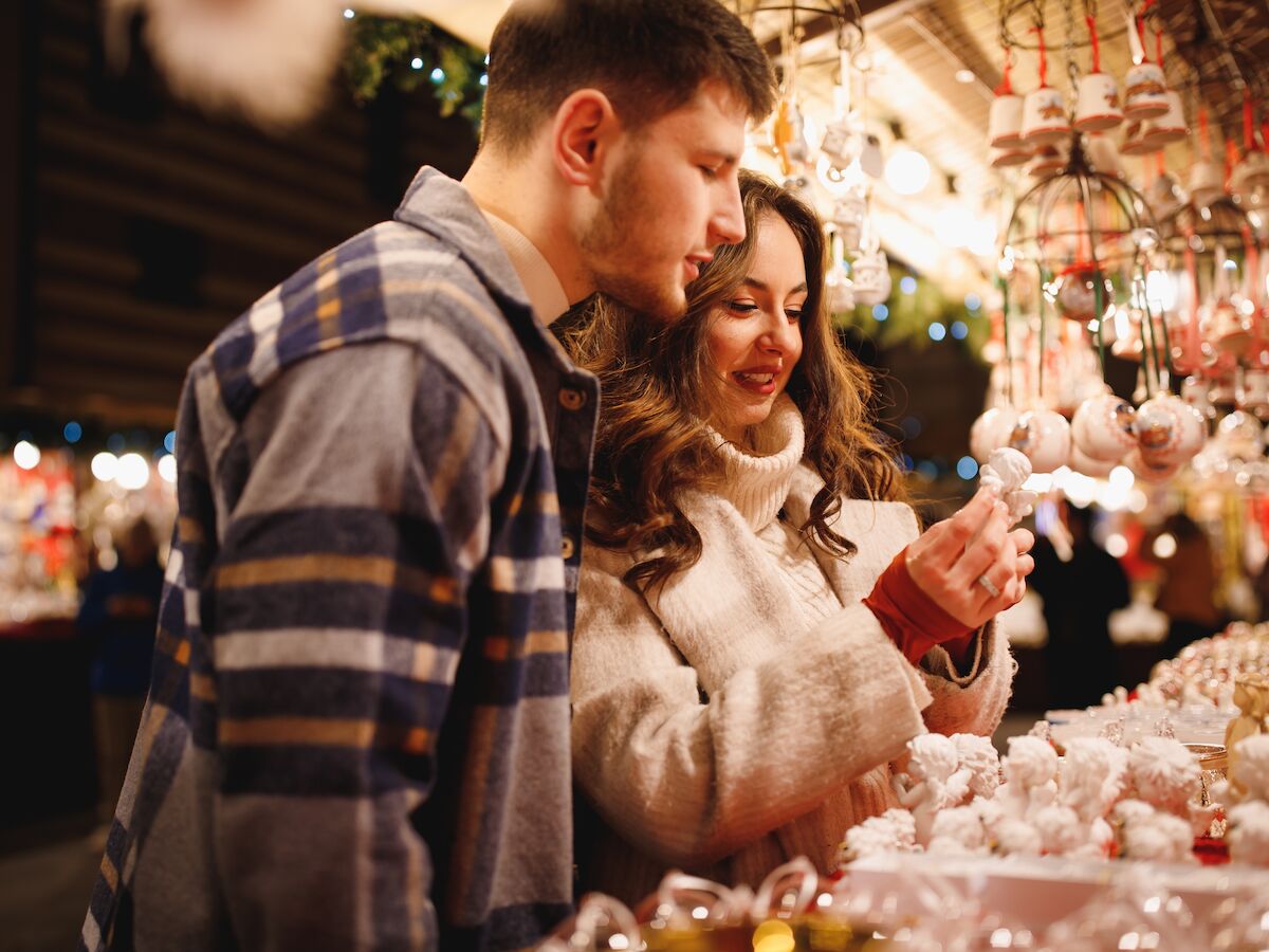 A couple browses a festive market, admiring and touching ornaments and lights at a holiday stall, warm glow around them.