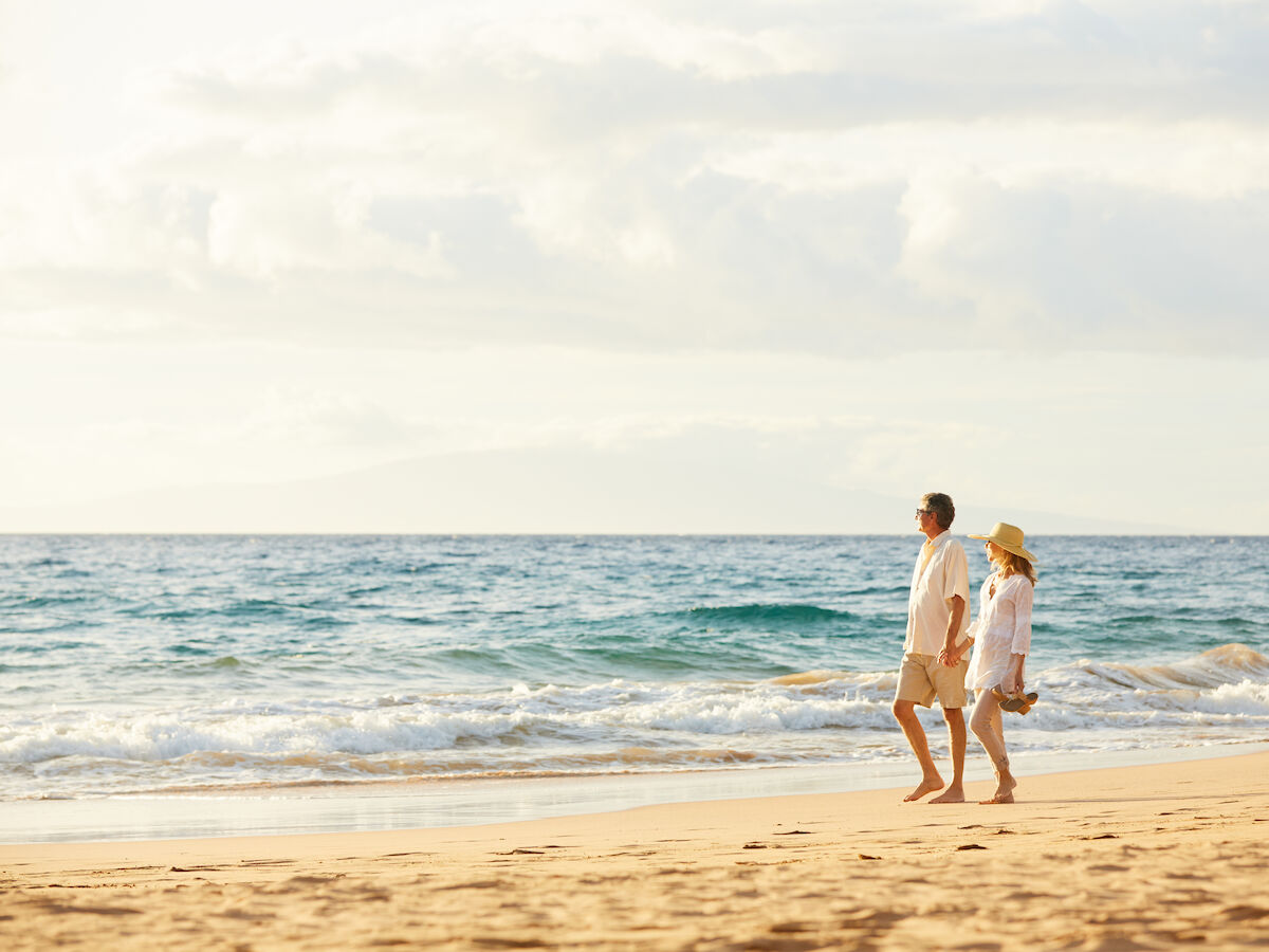 Two people walk arm-in-arm along a sunny beach, waves lick the shore, and a calm sea stretches to the horizon.