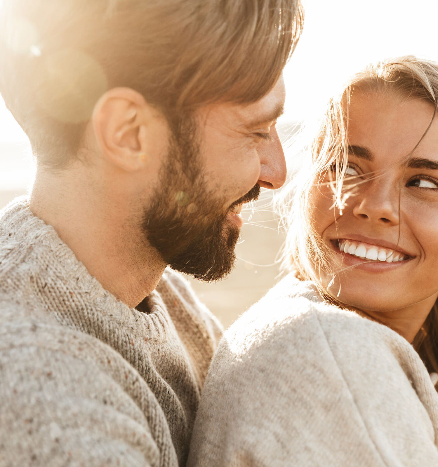 A couple is sitting together on a sunny day, looking at each other and smiling warmly, with a soft light in the background.
