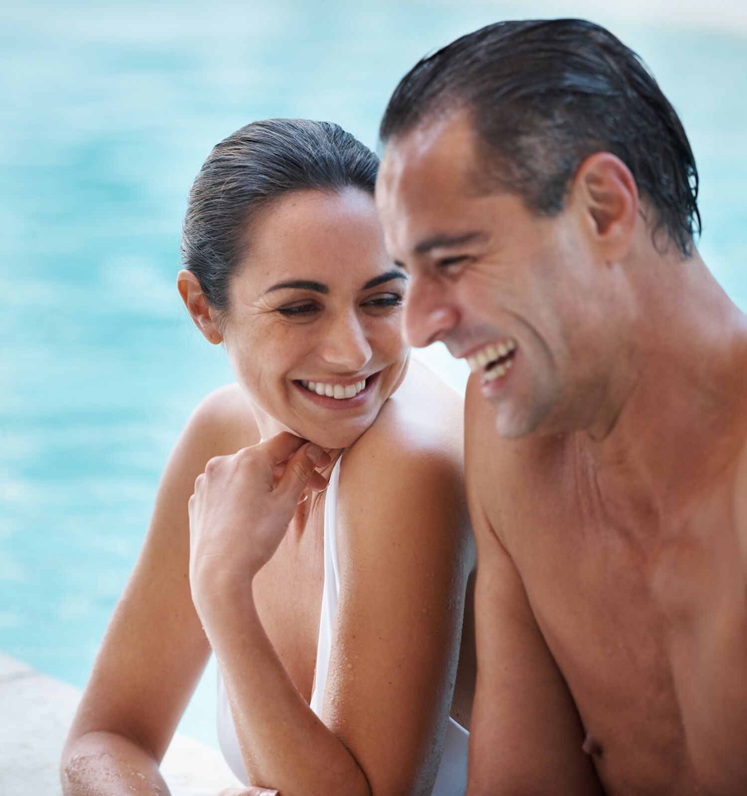 A man and woman are smiling and laughing while relaxing by a pool.