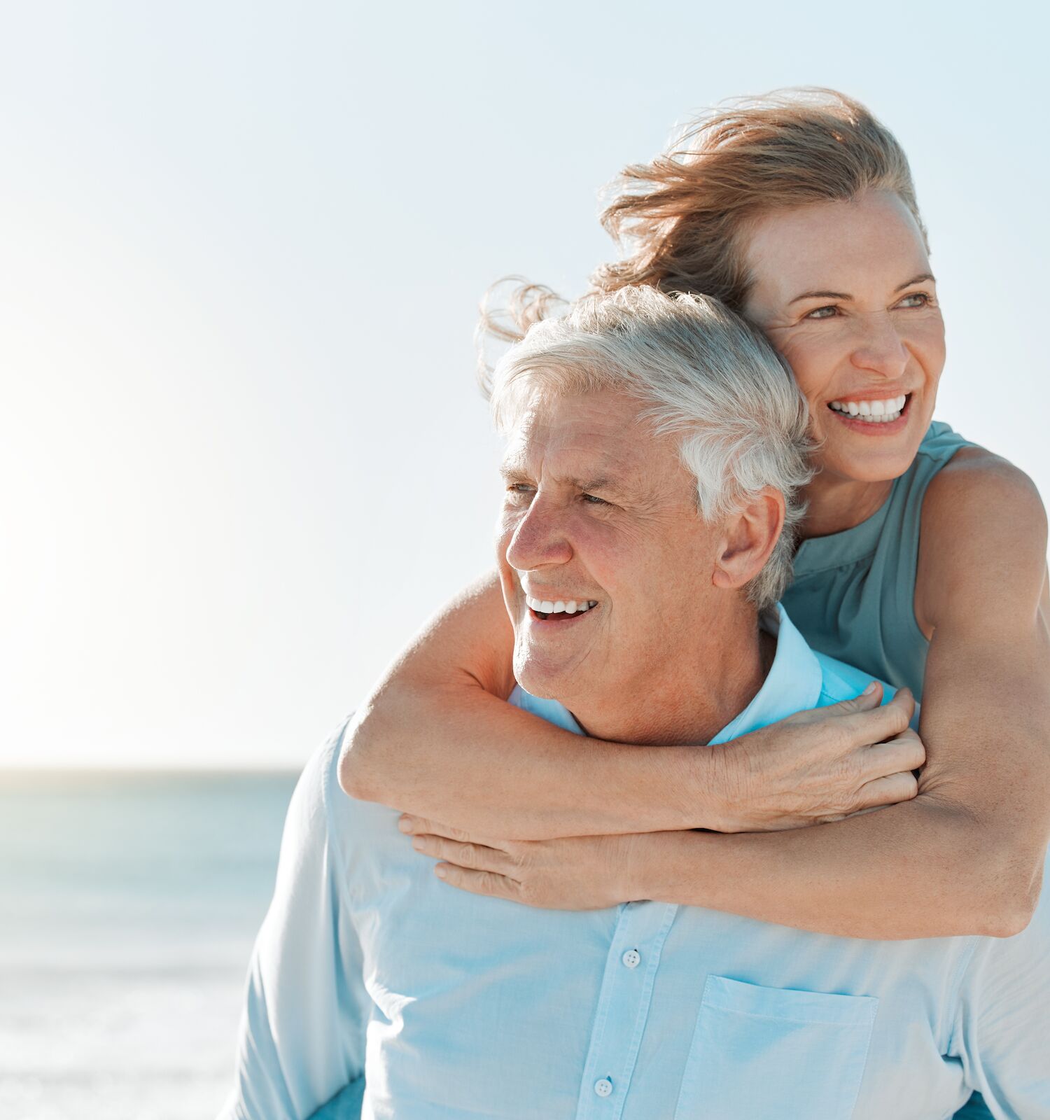 An older couple smiles at the beach, the woman embracing the man from behind, with the ocean in the background.