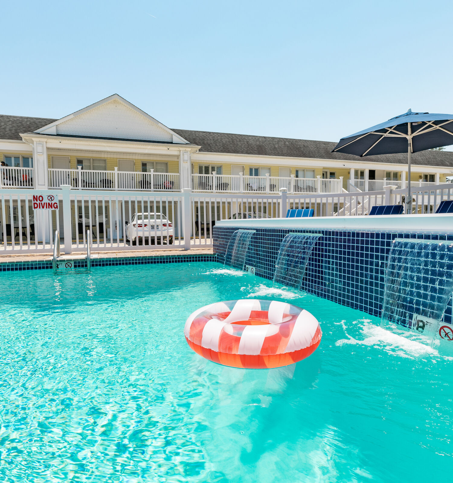 A pool with a striped float, surrounded by a fenced area. A building with balconies and patio umbrellas is in the background.
