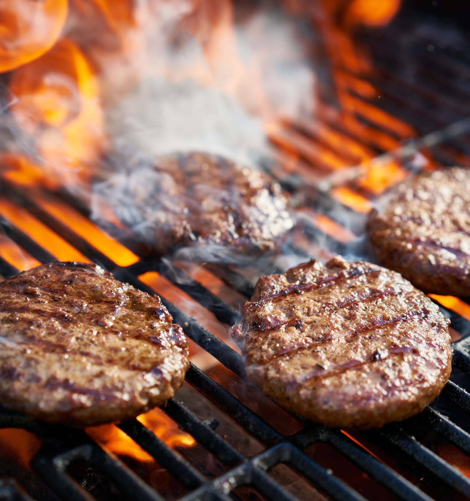 Four hamburger patties are grilling over open flames, with visible smoke rising on the barbecue grill.