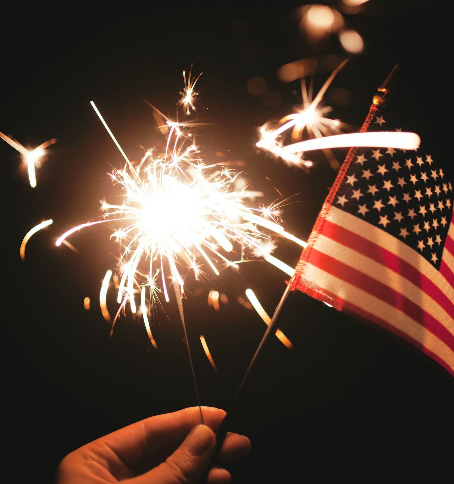 A hand holds a sparkler and a small American flag against a dark background, creating a festive and patriotic scene.