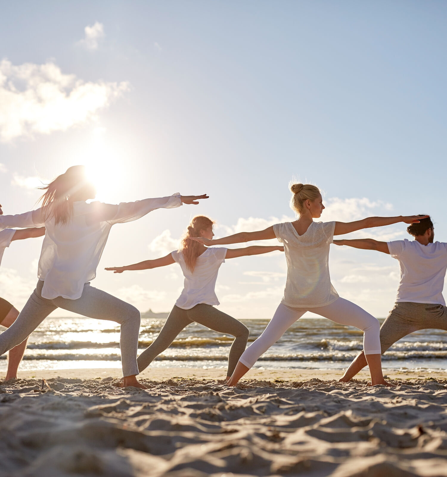 A group of people are practicing yoga on a beach, standing in a warrior pose, with the sun setting in the background.
