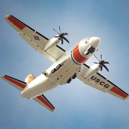 The image shows a US Coast Guard aircraft in flight, featuring distinctive USCG markings and a bright color scheme.