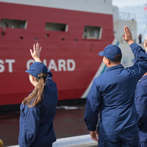 Three people in navy blue uniforms wave at a red and white Coast Guard ship.