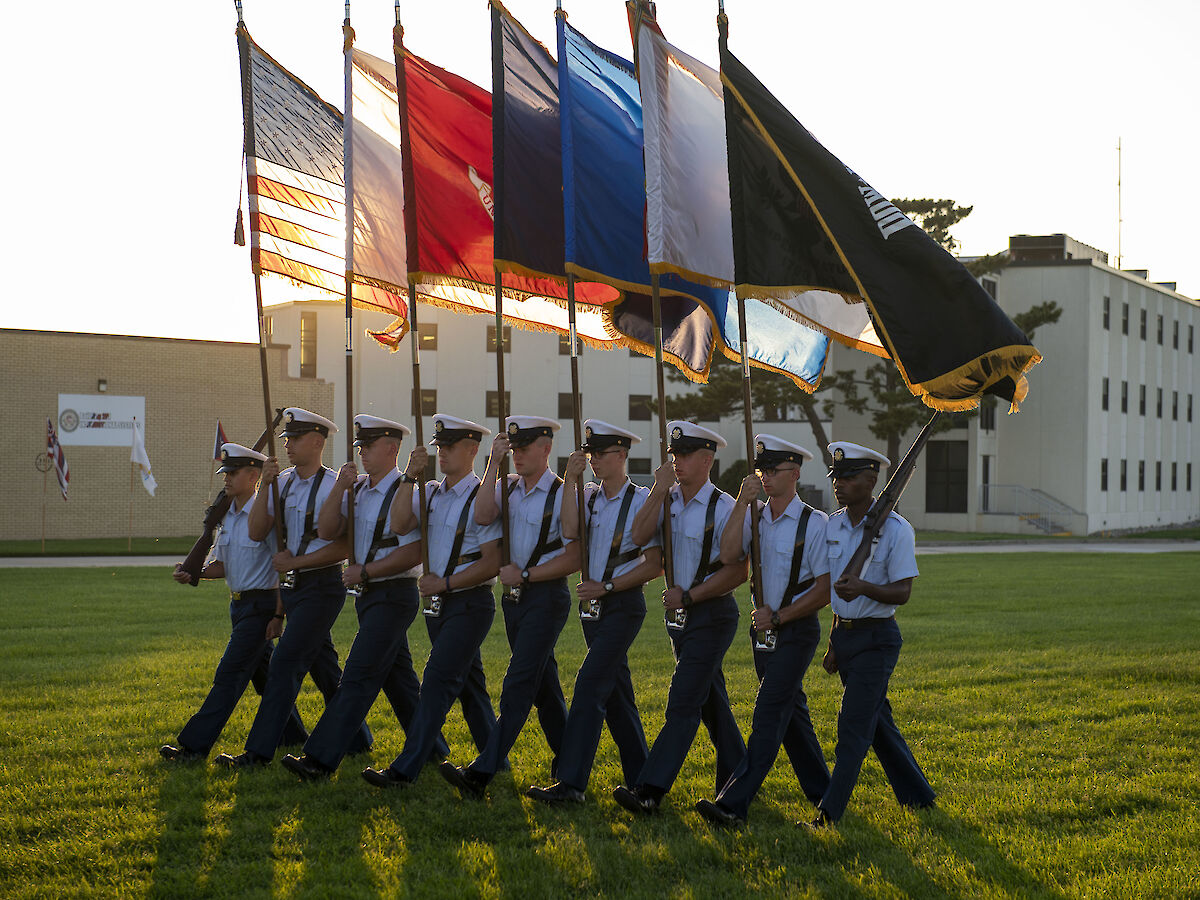 A group of uniformed individuals are marching in formation, each holding a different flag, on a grassy field beside buildings.