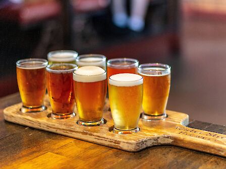 A wooden serving tray holds eight small glasses of different types of beer, placed on a wooden table in a cozy setting.