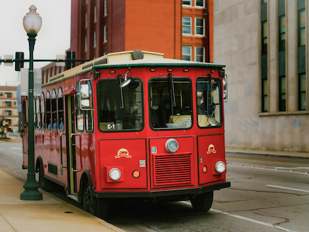 A red trolley bus is parked on a city street, surrounded by urban buildings and a streetlamp.