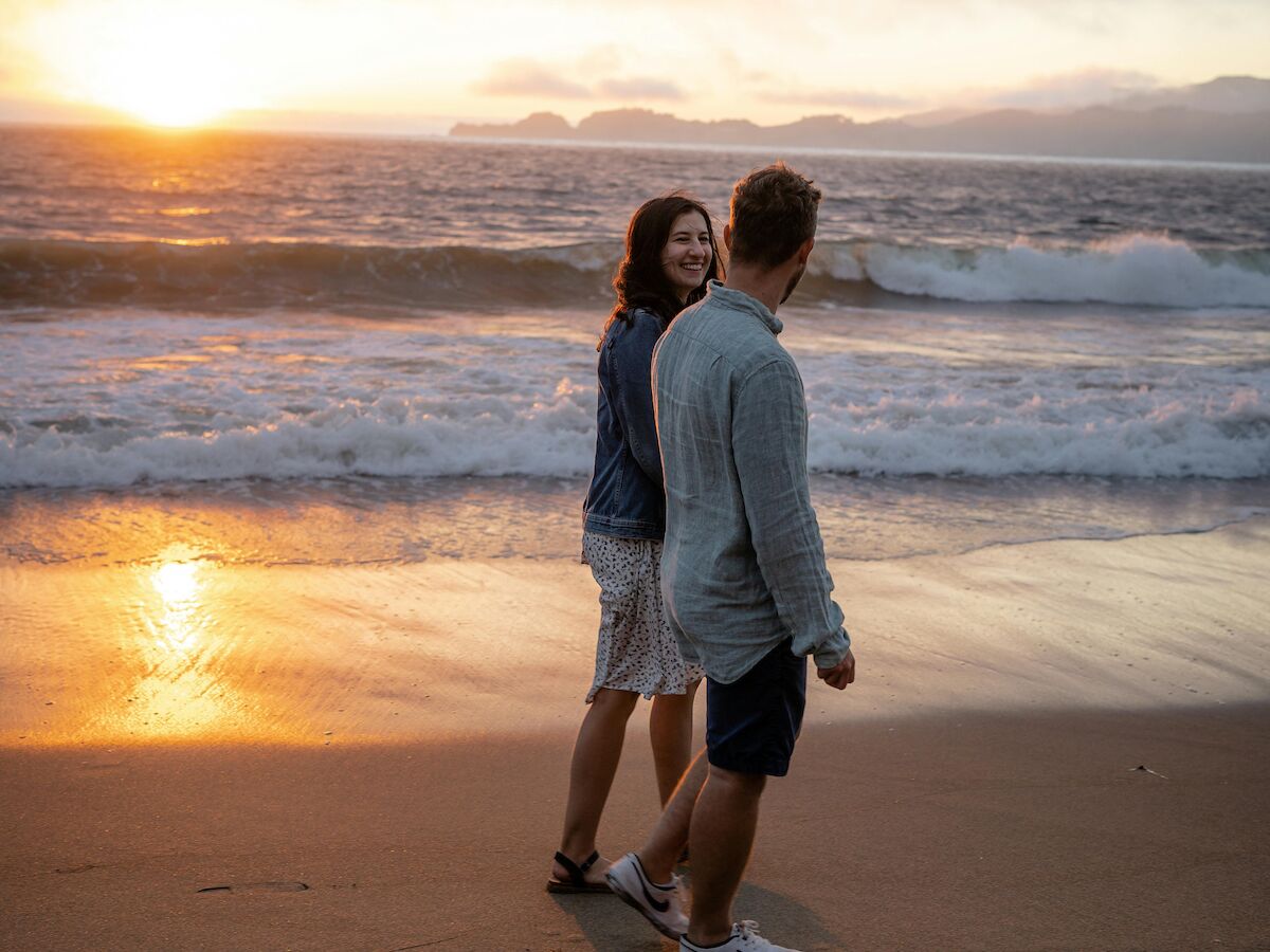 A couple is walking on the beach during a sunset, with waves gently crashing onto the shore.