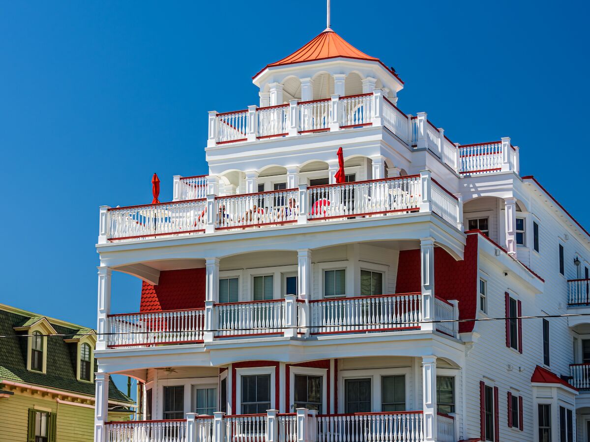 A white, multi-story coastal hotel with red accents and balconies, a red awning, landscaped front yard, and an American flag atop the roof.
