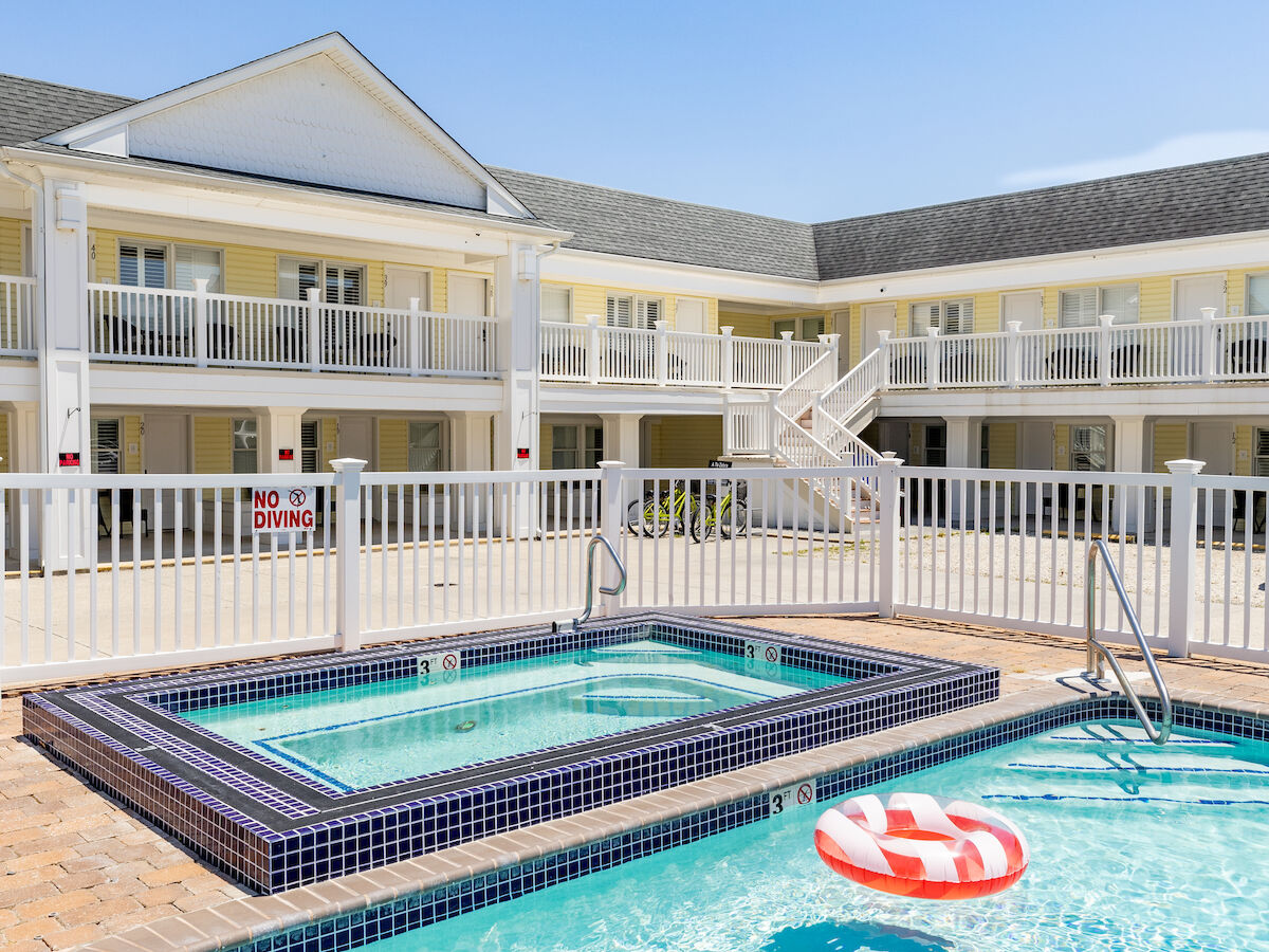 A bright hotel-style pool area with a rectangular main pool and a raised hot tub, surrounded by a white railing and a two-story cream building, under a clear blue sky.