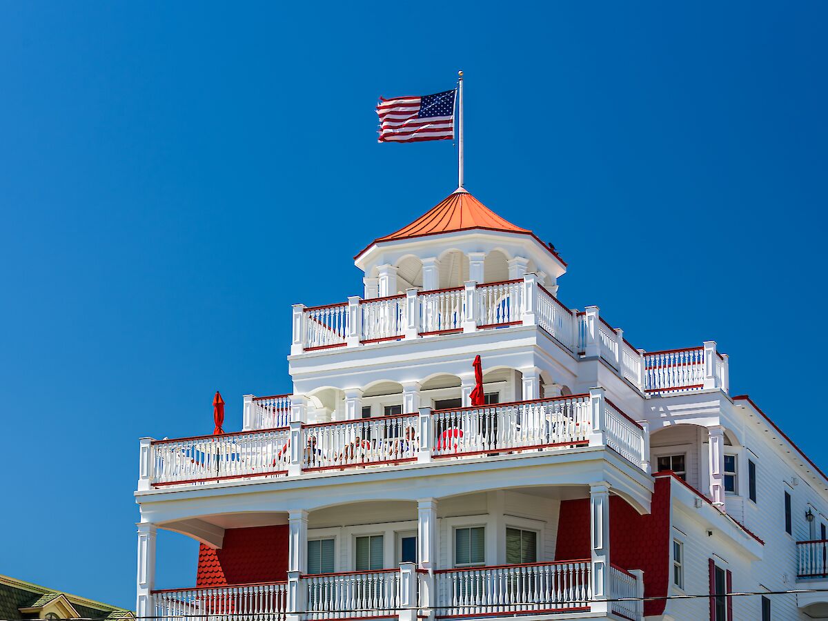 A white multi-story hotel with red awnings and railings, green lawn, shrubs, and a flagpole atop; bright blue sky overhead.