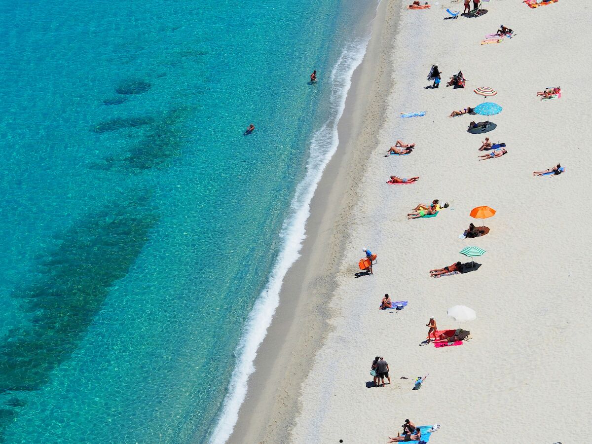 People sunbathing on a white sandy beach beside clear turquoise water, with colorful umbrellas and towels scattered along the shore.
