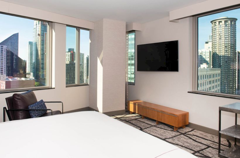 A modern room with a bed, chair, TV, and large windows showing a cityscape with tall buildings.