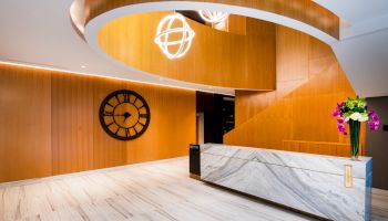 A modern, stylish lobby with a marble reception desk, a large wall clock, vibrant flowers, and a distinctive curved wooden staircase.