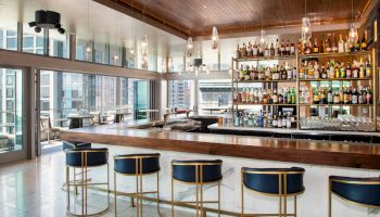 A modern bar with high stools, a well-stocked liquor shelf, large windows, and a city view outside. The space feels open and inviting.