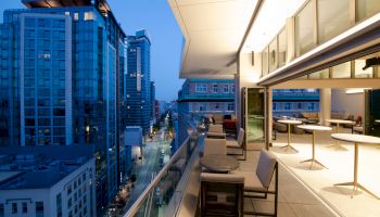 A modern urban balcony at dusk with seating and tables overlooks a city street lined with high-rise buildings and illuminated windows.