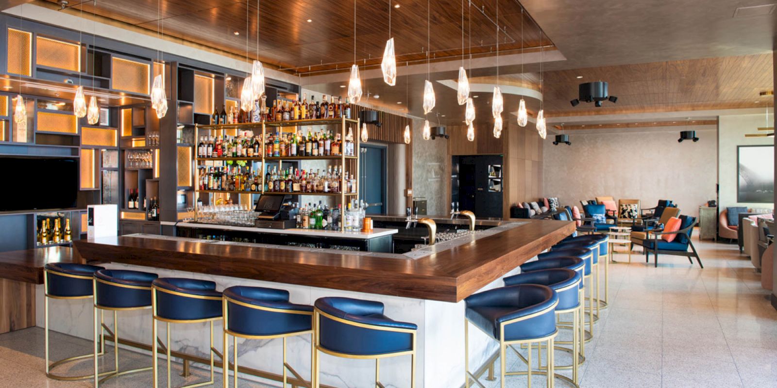 A modern bar with blue chairs, a long wooden counter, an array of hanging lights, and a well-stocked liquor shelf in a spacious, well-lit interior.