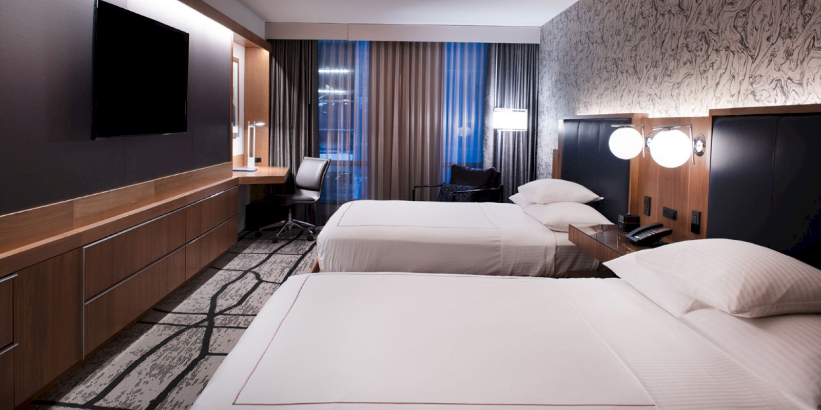 The image shows a modern hotel room with two beds, a large TV, a desk, and ambient lighting. The room has a contemporary design ending the sentence.