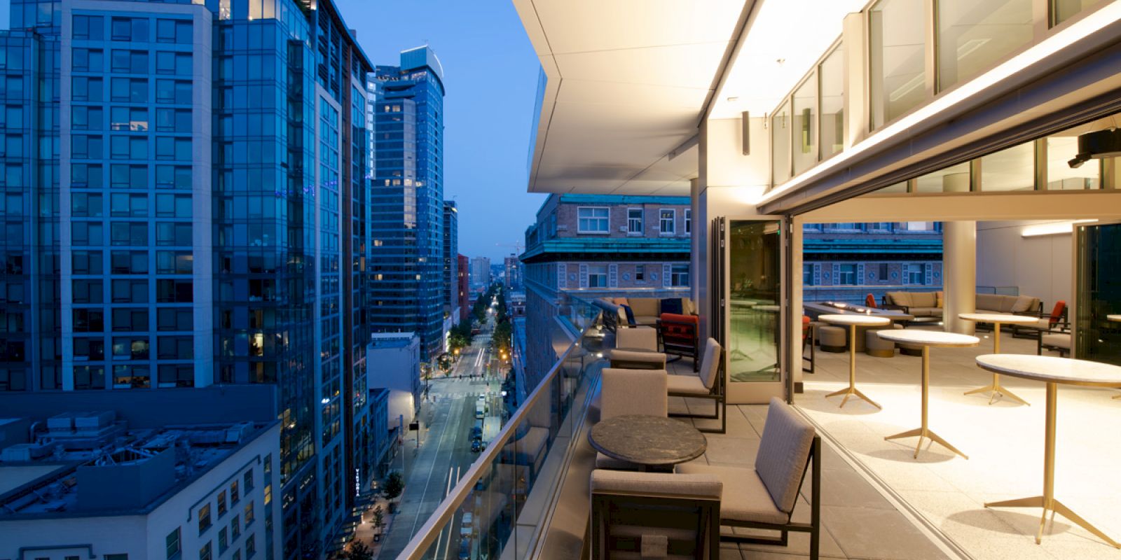 A modern balcony overlooks a cityscape at dusk, with buildings and a lit street below. The balcony has seating areas and tables.