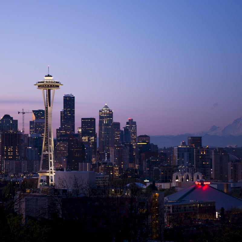 The image depicts the Seattle skyline at dusk, featuring the Space Needle prominently, with Mount Rainier visible in the background against a purple sky.
