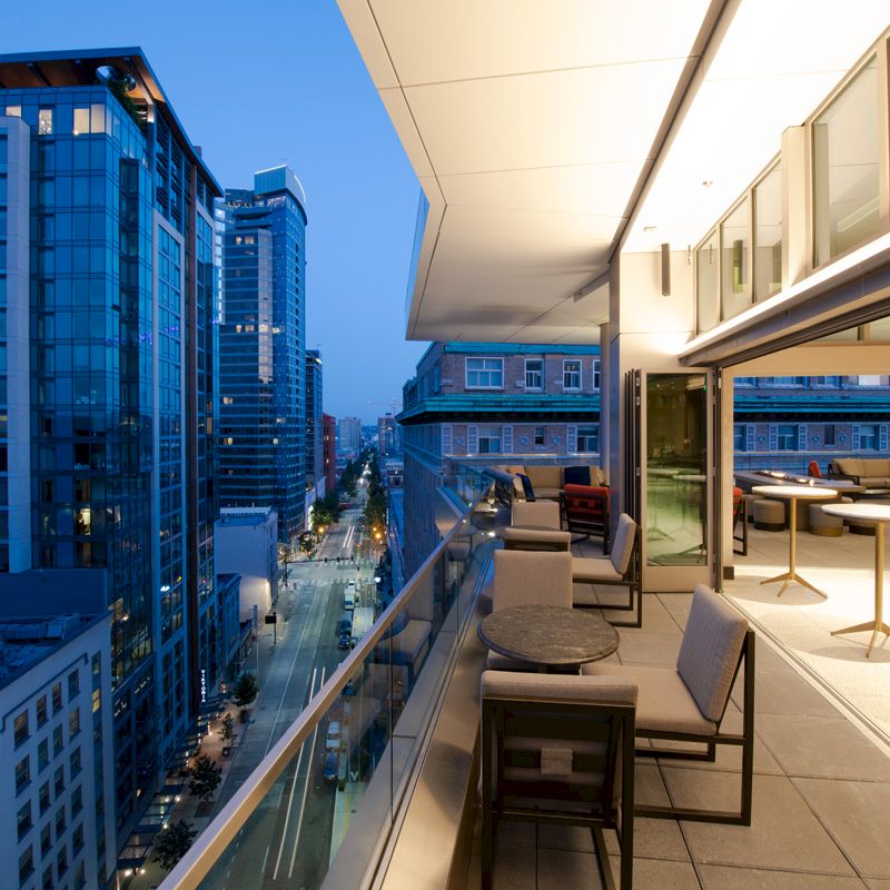 A modern rooftop patio with tables and chairs overlooks a city street with tall buildings at dusk, featuring a blend of glass and concrete architecture.