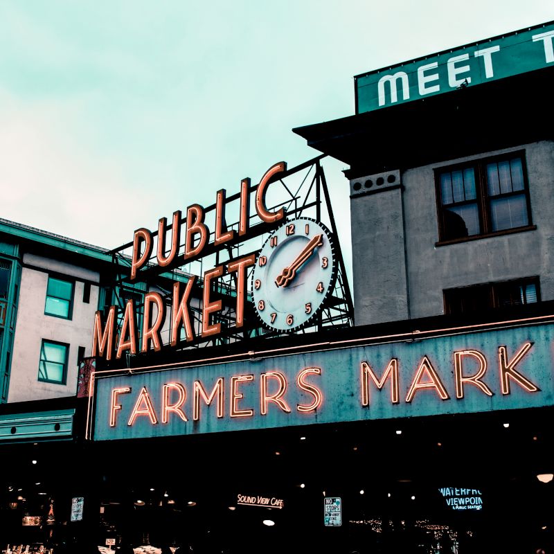 The image shows the iconic neon signage of a "Public Market" and "Farmers Market" with a clock, against a backdrop of buildings.