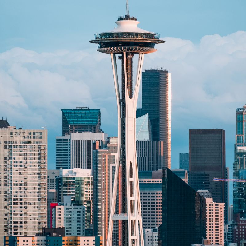 The image shows the Space Needle with a backdrop of buildings and skyscrapers under a partly cloudy sky.