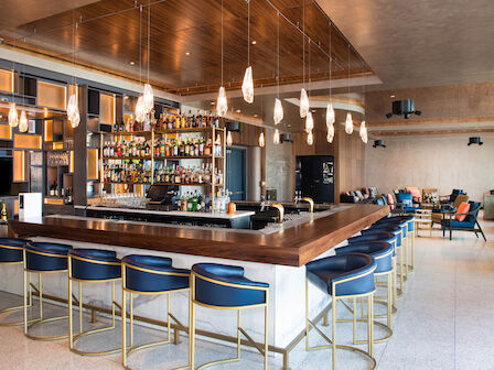 A modern bar with blue and gold stools, hanging lights, a well-stocked back bar, and a cozy lounge area in the background.