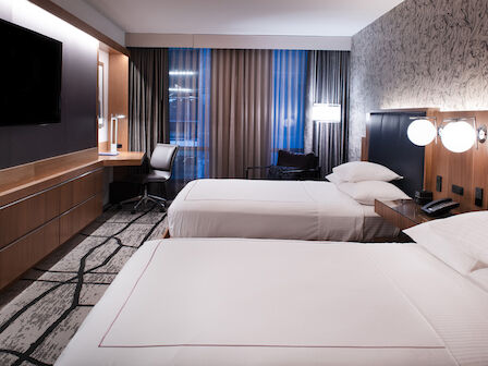 A modern hotel room with two neatly made beds, a desk with a chair, flat-screen TV, wall art, and contemporary lighting fixtures.
