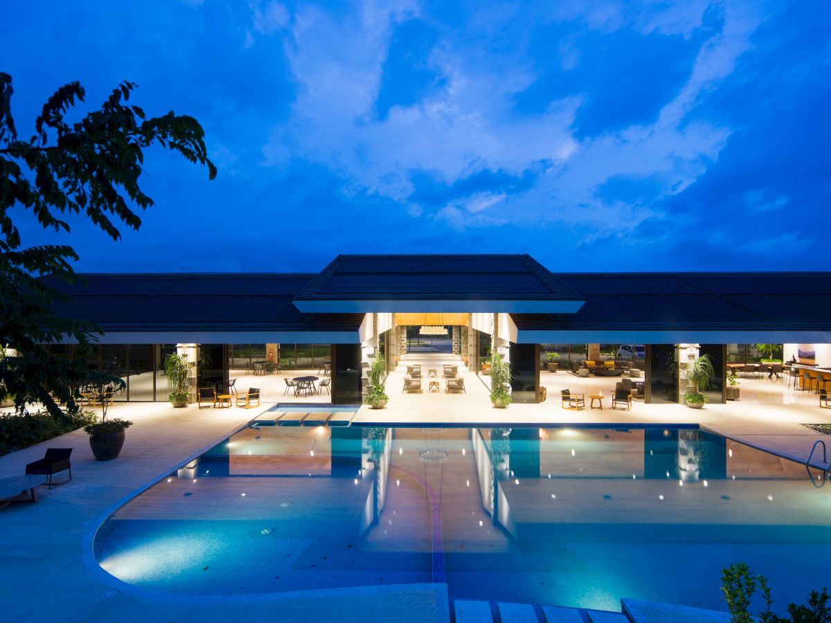 A modern, illuminated building with an open layout is visible at night, featuring a large swimming pool in the foreground and seating areas.