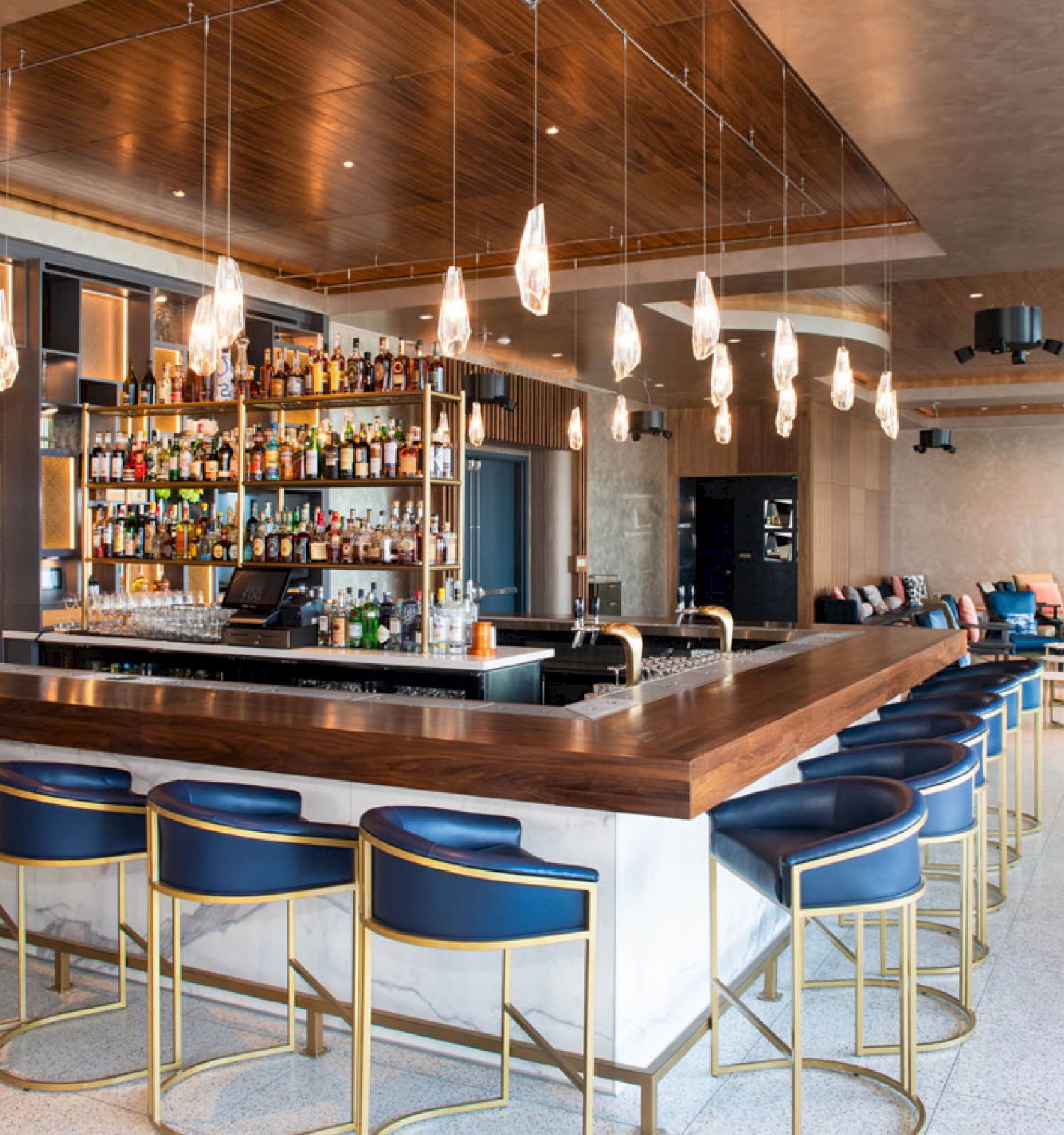 The image shows a modern bar with a wooden countertop, blue chairs, hanging lights, various bottles on shelves, and a seating area with couches.