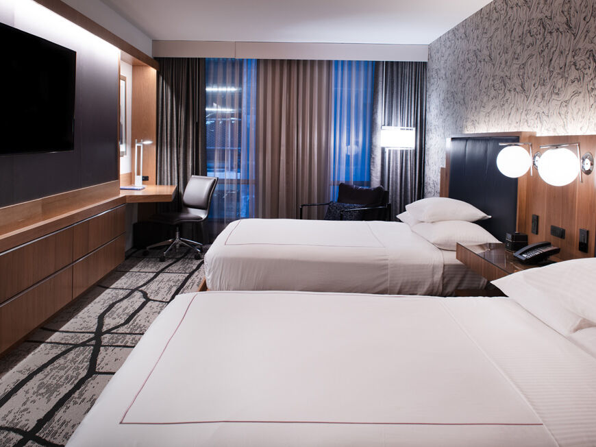 A modern hotel room with two beds, nightstands, lamps, a flat-screen TV, a desk, a chair, and large windows with curtains.