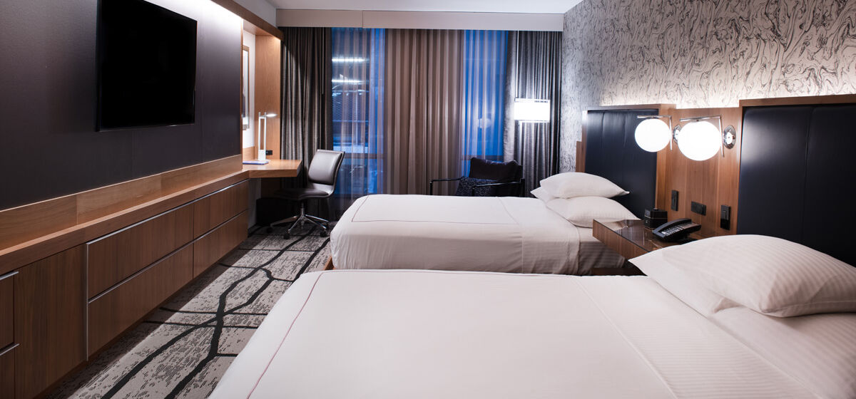 A modern hotel room with two beds, nightstands, lamps, a flat-screen TV, a desk, a chair, and large windows with curtains.