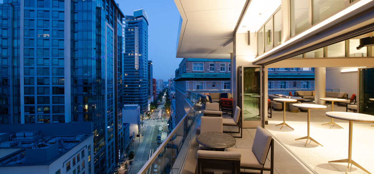 A modern cityscape viewed from a high balcony with outdoor seating and illuminated indoor area at dusk, surrounded by tall buildings and streets.