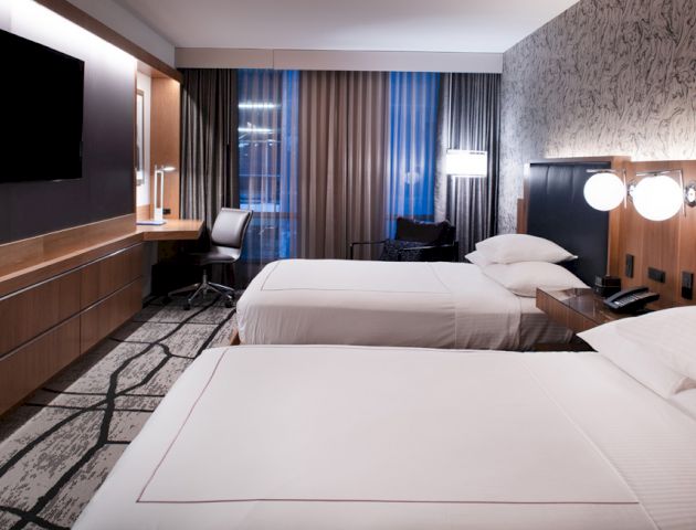 A modern hotel room with two double beds, a wall-mounted TV, a work desk with a chair, and contemporary decor including patterned wallpaper and lighting.