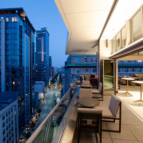 A modern balcony overlooking a city skyline at dusk, featuring seating areas and large windows into a building with tables inside.