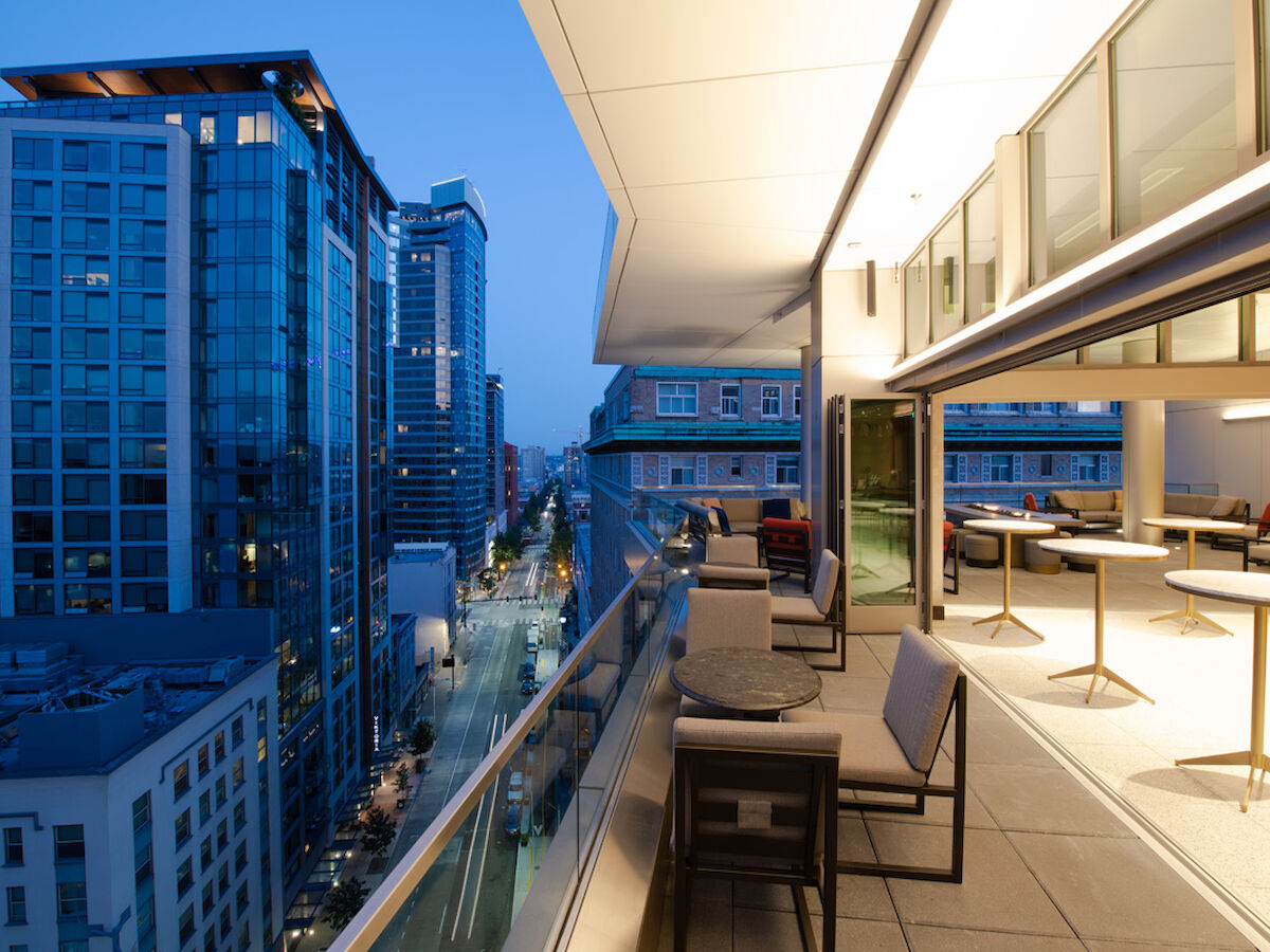 A modern balcony with furniture overlooks a street lined with tall buildings at twilight. The interior space features tables and tall stools.